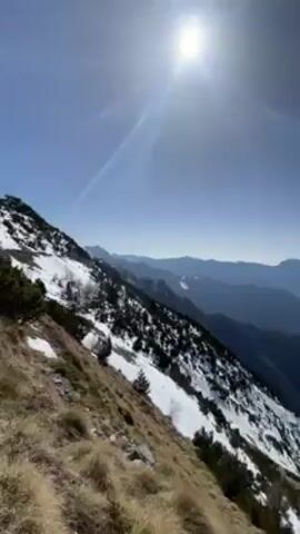 The video opens with a wide shot of a mountain range under a bright, sunny sky. The foreground shows a steep slope with a mix of dry, brown grass and patches of snow. As the camera pans, a valley comes into view between the mountains. The landscape includes distant peaks still covered in snow. Three people enter the frame, walking along the snow-covered ground. One of them has a large backpack. The camera continues to pan, revealing more snow and scattered trees.