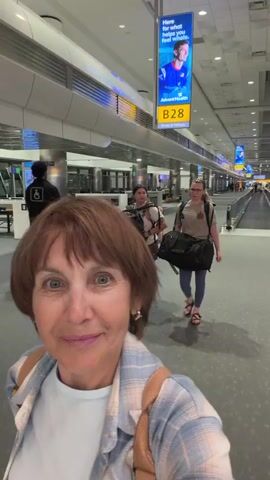 A woman with reddish-brown hair and blue eyes is filming herself in an airport terminal. She is wearing a light blue and white plaid shirt over a light blue t-shirt, with a tan backpack on her shoulders. She is smiling at the camera. Behind her, two other women are walking away from the camera down a long, brightly lit hallway. The hallway has a moving walkway on the right side. There are signs and displays along the walls. The woman in the foreground is centered in the frame, and the video appears to be a selfie.