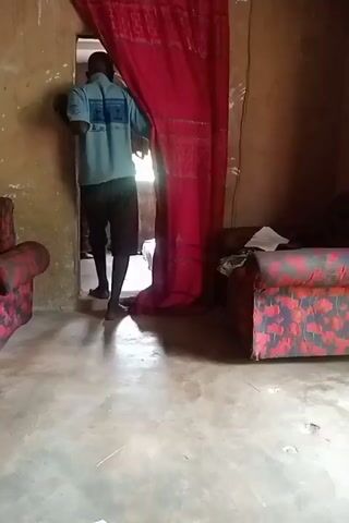 A man wearing a light blue t-shirt and dark patterned shorts walks through a doorway draped with a red curtain in a dimly lit room. The room has concrete floors and walls made of what appears to be mud or plaster. Two sofas, upholstered in a red and black floral pattern, are visible in the room. The man is walking from left to right, partially obscured by the curtain.