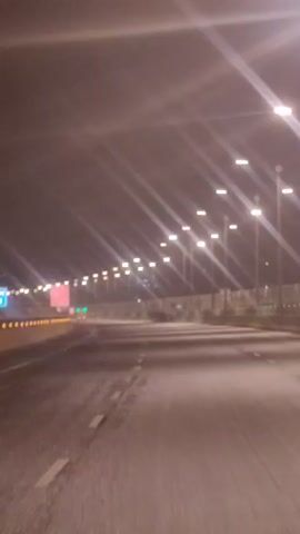 The video shows a nighttime view from a moving vehicle on a highway. The road is wet, suggesting recent rain. Streetlights illuminate the highway, creating streaks of light due to the camera's movement. A safety barrier lines the side of the road, with lights along its top. There are various signs visible, including a blue sign with "KM 12" and a red and white sign with text that is illegible due to the distance and motion blur. The overall atmosphere is dark and somewhat blurry, typical of footage captured from a moving vehicle at night.