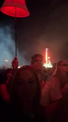 The video shows a group of people at night, apparently at a concert or festival. The lighting is dim, primarily red, and there is smoke in the air. In the foreground, a woman with long hair holds the camera, taking a selfie with a man standing behind her. The man is wearing sunglasses and a baseball cap. He's holding a drink in one hand. Behind them, there's a crowd of people, and in the distance, a structure is lit up with bright lights and possibly fire. Someone is holding a red lampshade on a stick above the crowd.