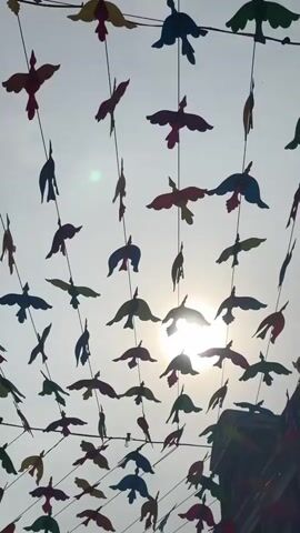 This is a low-angle shot looking up at a bright sky with the sun partially obscured by clouds and a line of colorful paper birds strung across overhead wires. The birds, in shades of red, yellow, blue, and green, are cut into silhouette shapes and are suspended by thin black strings, creating a festive, decorative display. Some birds are angled as if in flight, while others hang downwards. The background is a hazy, pale sky with hints of bright sunlight peeking through. The bottom right corner of the frame shows the upper facade of a building, indicating an outdoor, possibly urban, setting. There are no people visible in the video.