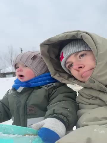A close-up shot shows a young adult and a toddler in a snowy environment. The adult, likely a parent or guardian, is on the right, wearing a tan hooded jacket and a gray knit hat with a red patch. The hood is up, partially obscuring their face. They look at the camera, initially with a slightly open mouth, then they smile. The toddler, on the left, wears a green winter jacket, a blue scarf, and a gray knit hat. The toddler's mouth is slightly open, and they appear to be looking off to the side. They are holding a light blue sled. The background is blurred but suggests a snowy field with bare trees and possibly a distant fence.