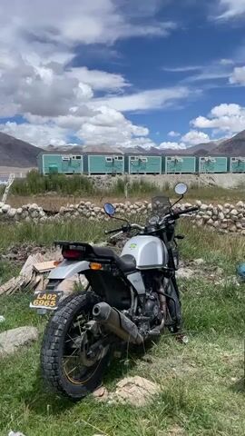 A motorcycle is parked in a grassy area with a stone wall in the foreground. In the background, there are teal-colored buildings lined up, mountains, a body of water, and a blue sky with white clouds. The motorcycle is facing away from the camera, with its rear wheel visible. The license plate on the motorcycle reads "LA02 6965". The video pans from the motorcycle to the wider landscape.