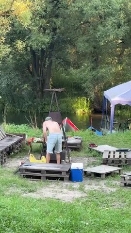 A man is standing in front of a grill outdoors near a body of water. He is wearing shorts and is shirtless. There are several wooden pallets scattered around the area, some serving as makeshift tables. A blue and white cooler is visible on one of the pallets. In the background, there is a body of water with trees lining the bank. A white tent is set up near the water, and some boats are visible in the water. Further in the background, a wooden structure with speakers stacked on it can be seen.