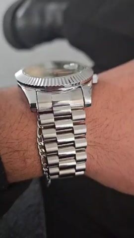 A close-up shot shows a silver-colored Nibosi wristwatch on a hairy wrist. The watch has a green face with silver hands and Roman numerals. The day is displayed as "Thursday" at the top of the face, and the date is shown at the three o'clock position. The watch has a fluted bezel and a jubilee-style bracelet. A black shoe is visible in the background. The camera stays fixed on the watch on the wrist.