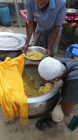 Two middle-aged men are outdoors preparing food. One man, wearing a blue polo shirt and a plaid sarong, holds a silver bowl filled with yellow food, transferring it into a large silver pot. The pot is nearly full of the same yellow substance. The second man, wearing a white patterned skullcap and a plaid sarong, uses a white spatula to mix the food in the pot. A large yellow cloth rests on the edge of the pot. A blue barrel and other pots and pans are visible in the background. The location seems to be in a courtyard or backyard, judging by the brick wall and partial views of a shed. A third man is visible in the background, standing near a small table covered with a red-and-white checkered cloth.