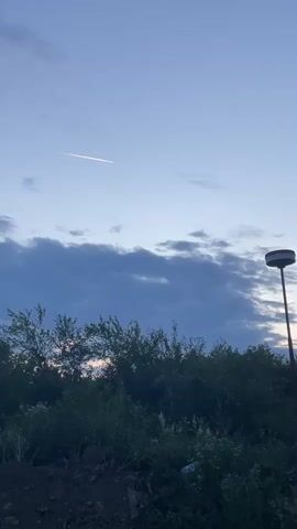 The video shows an outdoor scene at what appears to be dusk or dawn in Kinchesh, Ukraine. The sky is a light blue with scattered, darker clouds forming a band across the middle. A bright, white linear streak, possibly a contrail from an aircraft or a meteor trail, is visible in the upper left portion of the sky, extending horizontally. In the foreground, dense green bushes and foliage occupy the lower half of the frame. On the far right, a tall lamppost with a dark, circular light fixture stands, silhouetted against the sky. No people or visible text are present in the video.