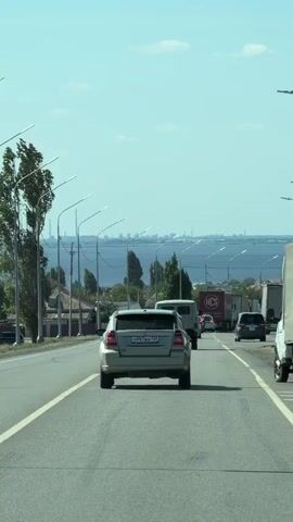 A gray car is driving on a paved road with a body of water in the background. The car is in the center of the frame, and the road has white lines marking the lanes. There are other vehicles on the road, including a white van, several trucks, and other cars. In the background, there is a clear sky with a few clouds. The body of water appears to be a lake or a sea, with some buildings and structures visible on the distant shore. Trees and streetlights line the side of the road. The video is shot from a moving vehicle, likely following the gray car.