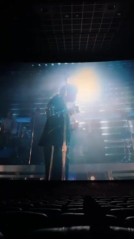 The video shows a person in a movie theater, watching a concert film of Michael Jackson on the big screen. Jackson, in a black jacket and pants, is seen moving on a stage bathed in spotlight, performing for a large audience. Other performers are on stage playing keyboards and drums. The theater seats are visible in the foreground, and the theater's ceiling is a dark, grid-like structure.