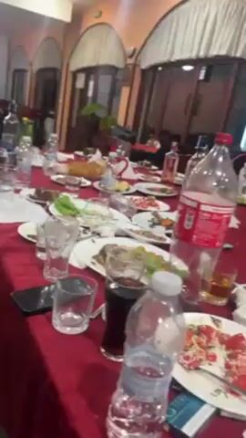 The video shows a messy dining table in what appears to be a restaurant or party venue. The table is covered with a red tablecloth and is littered with empty plates, glasses, water bottles, soda bottles, and cigarette packs. Several men are seen in the video: one is wearing a yellow shirt and looking at his phone, while others are near a door or walking around. The overall atmosphere suggests a gathering or celebration that has just ended, with the room in a state of disarray.