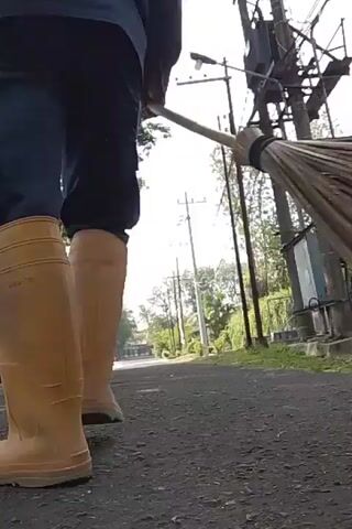 A person wearing a black shirt, black shorts, and tan boots is sweeping a dirt road with a broom made of natural fibers. The person is walking away from the camera, towards the right side of the frame. In the background, there are trees, utility poles with electrical equipment, and a building. The lighting suggests it is daytime. The video is shot from a low angle, close to the ground, looking up at the person and the environment.
