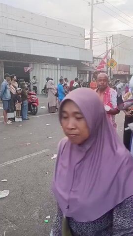 The video shows a street scene with several people. In the foreground, a person dressed as Ultraman walks towards the left, holding a wooden stick. To the left of the Ultraman character, another person in a white shirt stained with red, resembling blood, walks. This person is also holding a wooden stick. In the background, there are buildings, motorcycles, and a crowd of onlookers. Some people are wearing religious head coverings. A flag with red and white stripes, possibly a national flag, is visible. There is debris on the street. The overall impression is of a public event or gathering.
