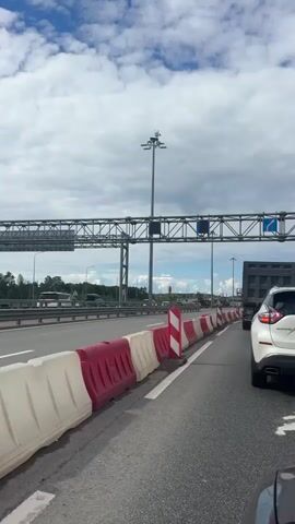 The video shows a view from the side of a highway, with a lane of traffic to the right and a barrier of red and white plastic water-filled traffic barriers separating it from another lane of traffic to the left. In the distance, there are trees and a cloudy blue sky. Overhead, a large metal structure spans the highway, likely a gantry for traffic signs or lights. A white SUV is visible in the right lane, and other vehicles can be seen further down the road. The traffic appears to be moving slowly or stopped, suggested by the presence of the barriers and the context of a highway. The lighting indicates it is daytime.