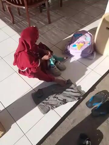 A young child, wearing a red hooded garment and a red hijab, sits on a white tiled floor. The child is wearing grey shoes and what appears to be a light blue brace or sock on their right leg. In front of the child is a pair of grey pants. To the right of the child is a purple backpack adorned with an ice cream cone graphic. Behind the child is a wooden chair and part of a table. Sunlight casts shadows across the floor.