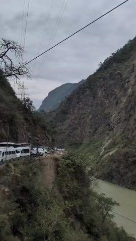 This video captures a scenic view of a river flowing through a mountainous region. The river has a murky, greenish-brown color and is surrounded by steep, rocky slopes covered in patches of green vegetation. On the left side of the frame, a road is visible, with several vehicles parked along its edge. The sky is overcast, contributing to a muted lighting condition. The camera appears to be moving, offering a panoramic perspective of the landscape, including a prominent power line spanning across the frame. The overall impression is one of a remote and rugged natural environment.