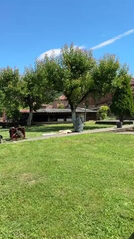 The video depicts a sunny outdoor scene with lush green grass, trees, and gardening tools. In the foreground, there are two black objects, one round and one rectangular, along with a coil of white rope. In the midground, a white dog is resting peacefully on the grass, observing its surroundings. Behind the dog, there is a metallic structure that resembles a greenhouse or a large arbor, with green netting covering its top. In the background, there are buildings with red roofs, suggesting a rural or village setting. The sky is clear and blue, with a few white clouds. The camera slowly pans from left to right, capturing the details of the environment.