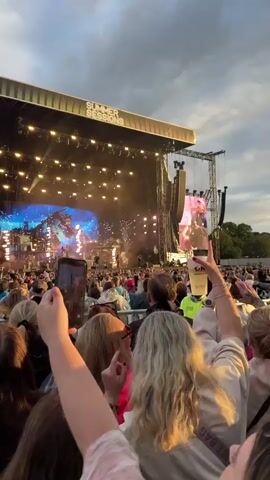 The video shows a concert crowd facing a large stage with bright lights and a screen displaying performers. Several people in the foreground are holding up their phones, some with flashlights on, to record the event. One person in the middle ground is wearing a bright yellow safety vest. The sky above is partly cloudy. The overall atmosphere appears to be one of excitement and engagement with the performance.