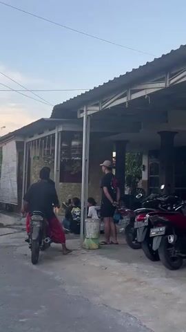 The video shows a group of people outside a building. In the foreground, a person is sitting on a motorcycle. They are wearing a red sarong and a black shirt. Another person, wearing a black t-shirt and shorts, is standing to the right of the motorcycle, facing away from the camera. Behind them, two more people are sitting on the ground. One person is wearing a black t-shirt with a graphic on it, and the other is wearing a black shirt and shorts. The building has a covered outdoor area with a metal roof supported by pillars. There are motorcycles parked in front of the building. The environment appears to be an outdoor residential area or a commercial space with a rustic feel. There are some wires and cables on the ground, suggesting some sort of setup or event. The lighting suggests it is either early morning or late afternoon.
