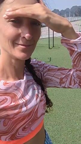 A woman wearing a patterned long-sleeved crop top and printed leggings is standing on an outdoor exercise machine on a bright, sunny day. She shields her eyes with her hand, smiles, and appears to be recording a video. The exercise machine is green and shows signs of rust. The ground is covered with artificial turf, and palm trees line the background. A view of a bay is visible in the distance.