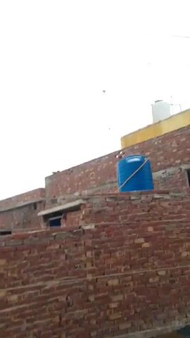 A cloudy sky is visible above a series of brick buildings. Three parallel wires run horizontally across the frame. On the roofs of the buildings are water tanks, one white and one blue. A green tarp is draped over a section of one of the buildings. Below the buildings, a sign with the word "PEPSI" is visible. The video appears to be filmed from a low angle, looking upwards at the buildings and the sky. The overall impression is one of an urban or semi-urban environment on an overcast day.
