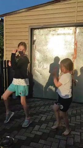 Two young girls are dancing in front of a building with a corrugated metal door. The girl on the left is wearing a black hoodie, a white shirt, and teal shorts. She has blonde hair tied in a ponytail. The girl on the right is wearing a white t-shirt and black shorts. She has brown hair. They are both barefoot except for the girl on the left, who is wearing sneakers. The ground is paved with brick-like stones. The sun is setting, casting long shadows.