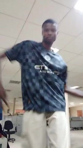 A slender, dark-skinned man dances in a large office space. He wears a dark gray t-shirt with "ETIHAD AIRWAYS" printed across the chest and light beige pants. The room has a tiled ceiling with recessed fluorescent lights. An office chair and metal filing cabinets are visible in the background, and other office equipment can be seen in the distance. The man moves his arms and shifts his weight from side to side as he dances.