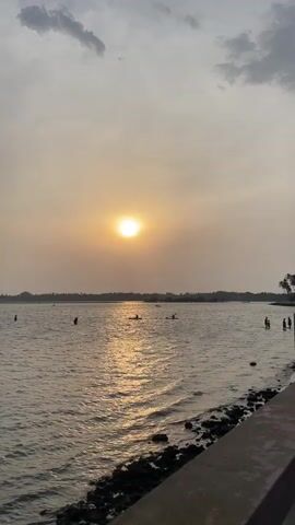 The video captures a sunset scene over a body of water, possibly a lake or a wide river. The sun is a bright, hazy orb in the sky, reflecting intensely on the water's surface, creating a shimmering effect. Silhouetted figures of people are visible at various points in the water; some are standing near the shore, while others are paddling in kayaks further out. The shoreline is rocky with a concrete edge. A line of trees is visible in the distance. The sky is overcast with light gray clouds.