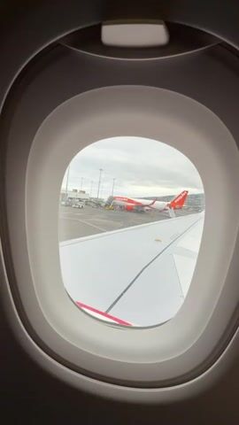 The video shows a view from an airplane window, revealing an EasyJet airplane parked on the tarmac. The plane has a white body with orange accents and the EasyJet logo. A stairway is connected to the plane's entrance, and several workers are visible around the aircraft and on the ground. The overcast sky suggests a cloudy day. The airplane's wing is visible in the foreground.