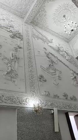A video captures the interior of a building adorned with elaborate white stucco reliefs on its walls and ceiling. The reliefs depict numerous figures in various poses, seemingly engaged in storytelling or religious narratives. Intricate patterns and ornamental borders frame the detailed scenes. Chandeliers hang from the ceiling, casting light on the ornate decorations. The camera pans across the expansive walls, revealing the scale and complexity of the artwork. The lower portion of the walls shows textured surfaces, possibly brickwork, beneath the stucco reliefs. An open doorway is visible at the bottom of the frame, offering a glimpse of the outside world where a tall, slender structure, possibly a water tower, stands against a bright sky. The overall impression is one of grandeur and artistic craftsmanship within a sacred or cultural space.