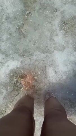 The video shows a pair of bare feet standing in shallow, clear water over a rocky, pebble-strewn seabed. The water is gently lapping around the ankles and feet. The sun appears to be shining brightly, creating reflections on the water's surface. The perspective is from above, looking down at the feet and the water.