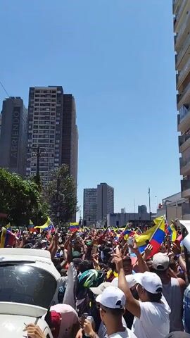 The video shows a large crowd of people gathered on a city street, many waving Venezuelan flags. The crowd is dense, and the people are dressed in casual clothing, with many wearing hats and sunglasses. Some are shirtless.  A few cars are visible, one white car in the foreground is crowded by people. Tall buildings line the street on both sides. The sky is clear and blue, suggesting a sunny day. The general atmosphere appears to be celebratory or protest-oriented.