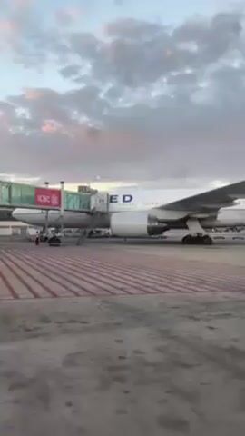 The video shows a large United Airlines airplane parked at an airport gate, connected to a jet bridge. The plane is white with a blue tail featuring the United globe logo. A couple of ground crew members are working near the front of the plane. The sky is cloudy with hints of pink and blue from the setting or rising sun. Other planes are visible in the distance. The camera moves slightly, focusing on the rear of the plane.