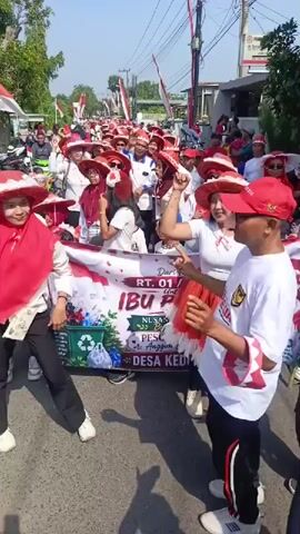 The video shows a group of people marching down a street, many of them wearing red and white clothing and hats. They are holding a banner that reads "IBU PERTIWI PESONA PIALA NUSANTARA LESTARI KEDUNGGALUR". Some people are dancing and waving. The street is lined with houses and trees, and there are flags visible. The overall atmosphere appears festive and celebratory.