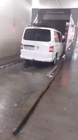 A white Volkswagen van with a roof rack is being moved through an automatic car wash. The vehicle is on a track, inside a well-lit indoor space. The car wash equipment, marked "WashTec," is visible around the van. The floor is wet, and there are metal tracks guiding the vehicle. The van's license plate is partially visible and reads "AUR SB 77". The car wash appears to be in operation, but no specific washing functions, such as spraying water or rotating brushes, are visible in the frame.