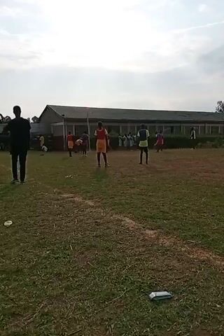 This video captures a group of young people engaged in a game of soccer on a grassy field. Several individuals, some wearing orange jerseys and shorts, are scattered across the field, appearing to be participating in the game. One person, dressed in black, stands on the left side of the frame, observing the activity. In the background, a long building with numerous windows suggests a school or similar institution. The sky above is partly cloudy, indicating it is daytime. The overall environment is an outdoor sports field.