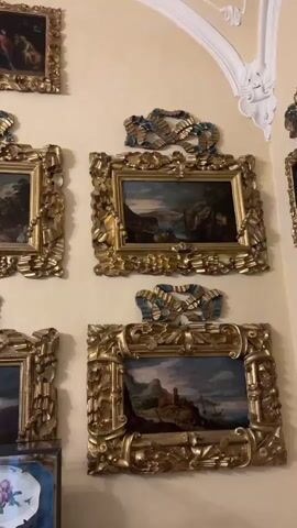 The video pans across a room adorned with numerous framed paintings, predominantly featuring ornate gold frames with decorative blue ribbon accents. The walls are a warm yellow, and the ceiling exhibits intricate white plasterwork, including a large chandelier. Several paintings depict landscapes and possibly historical or religious scenes. In the foreground, a person is partially visible, holding up a phone to take a picture or video of the artwork. Another person is also partially visible in the lower left corner. The overall impression is that of a museum, gallery, or grand interior space showcasing a collection of art.