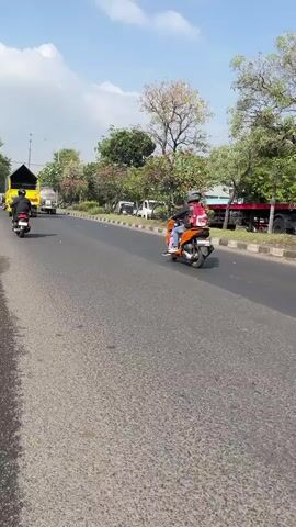 The media file captures a daytime scene on a paved road, likely in Gelam, East Java. The sky is bright with scattered clouds, suggesting fair weather. Two individuals are seen riding on motorcycles. In the foreground, an orange scooter is carrying two people. The rider wears a black helmet and an orange jacket, while the passenger, positioned behind the rider, has on a pink backpack and appears to be wearing a dark top and blue pants. The scooter is moving along the right side of the road. Further ahead and to the left, another motorcycle, ridden by a single person in black attire and a helmet, travels in the same direction.

The road is bordered by a grassy median with some trees and shrubs. Beyond the median, other vehicles are visible, including a yellow truck and what appears to be a red truck in the distance, along with smaller white vehicles. The overall setting depicts a typical urban or semi-urban environment with vehicular traffic. The lighting indicates it is likely late morning or early afternoon due to the bright sun and clear shadows. There is no visible text in the media file.