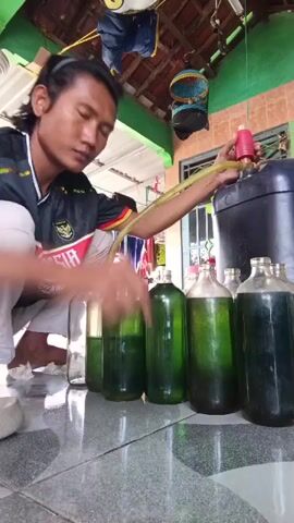 A man kneels on a tiled floor, holding a red manual siphon pump. He is transferring a green liquid from a large black plastic container into several clear glass bottles using a yellow hose. The man is wearing a black t-shirt with red and white accents and white pants. There are at least seven glass bottles filled with the green liquid lined up in front of him, and one empty bottle to his left. The background shows a thatched roof structure with green beams. Two bird cages are hanging from the ceiling. The lighting suggests it is daytime.