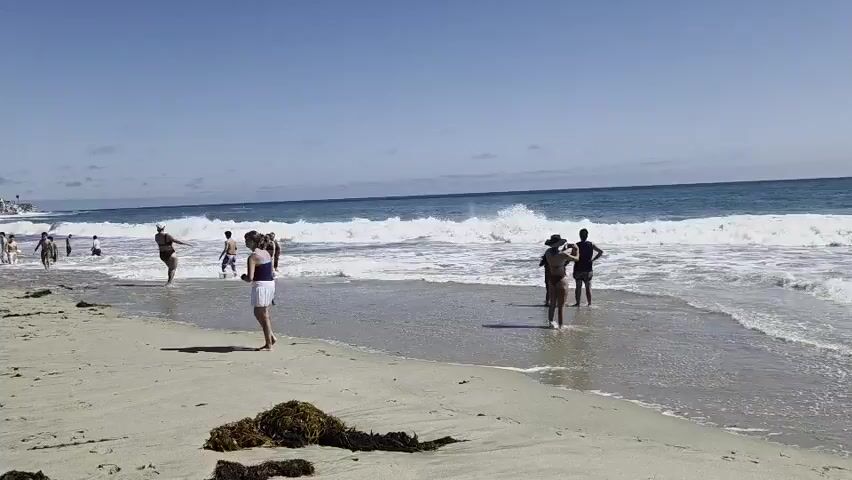 The video shows a bright, sunny day at a crowded beach. The sand is light colored with some patches of seaweed. In the background, there are gentle waves breaking on the shore. Several people are visible on the beach and in the shallow water. On the left side of the frame, a group of people are walking along the water's edge, and a few people are sitting on the sand. Closer to the center, a woman in a white skirt and blue top is walking away from the camera towards the water. Further out in the water, several people are standing and playing in the waves. On the right side of the frame, two people are standing in the shallow water, one of them appears to be holding a phone. In the far background, buildings and palm trees are visible on a hillside overlooking the beach. The sky is clear and blue with a few scattered clouds.