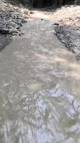 The video shows a muddy stream flowing through a narrow gorge with steep, rocky walls on either side. The water is flowing and appears to have a light brown or beige color. The gorge is lined with loose rocks and dirt, and there is some vegetation growing on the upper parts of the gorge walls. The sun is shining, casting light and shadows into the gorge, creating a dramatic effect. The overall impression is one of a wild and untamed natural environment.