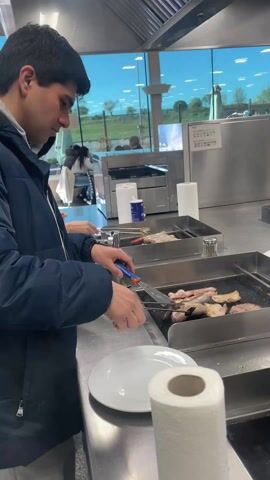 A man with dark hair is grilling various meats and mushrooms on two separate grill stations in a commercial kitchen. He is wearing a navy blue jacket and uses a spatula to handle the food. He pauses to smile at the camera briefly. Behind him is a large window showing an outdoor view of a field and other people. In the background, there is an industrial-style exhaust hood above the grills, along with other kitchen appliances and supplies, including paper towels, condiment shakers, and plates.