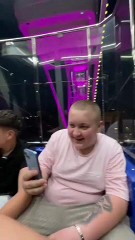 Inside a Ferris wheel car with mirrored panels, three young men are visible. One is holding onto a white pole, appearing excited, while the other is smiling, holding the pole as well. Another man, in a pink shirt, is seen holding a smartphone, smiling. The Ferris wheel car is lit with blue and white lights. The background shows city lights at night.