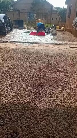 A person is using a red rectangular object to scrub a wet, paved surface. The person is bent over and appears to be exerting effort. There is a black car to the left, and various containers and equipment in the background. The ground in the foreground is covered in small, light-brown stones. The scene appears to be outdoors, possibly a driveway or yard, with buildings visible in the background. The lighting suggests it is daytime.