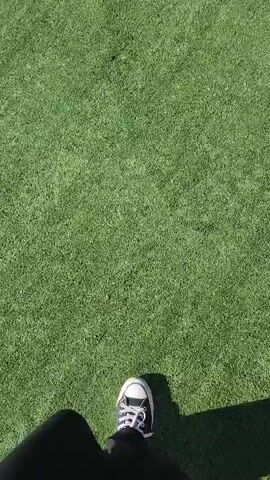 The video shows a person walking on artificial grass towards a small pond. The person is wearing black pants and Converse sneakers. The artificial grass is bright green, and the pond water appears greenish. The camera is positioned at the person's eye level, capturing their forward movement. The pond has a concrete edge and some greenery. The video appears to be taken during a sunny day as indicated by the sharp shadow visible on the ground.
