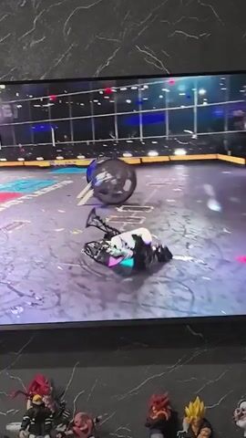 The video shows a robot combat competition on a screen. Two robots are visible in the arena. One robot is on its side with its wheels spinning, and the other robot is in motion, possibly attacking or maneuvering. The arena appears to be a circular or oval-shaped space with a textured floor marked with various colored squares and lines. The background of the arena includes glass walls enclosing spectators and possibly team members. The lighting in the arena is bright, with spotlights visible overhead. Below the screen, several anime figurines are displayed on a shelf.