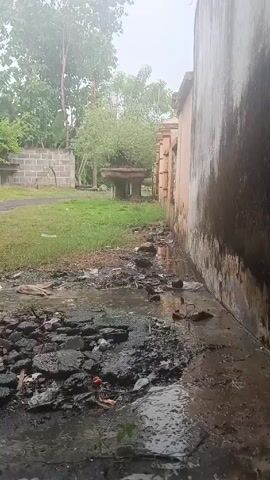 The video shows a muddy, garbage-strewn area next to a building on a cloudy day. 

The foreground of the video consists of a wet, cracked, and broken surface covered with various debris, including what appears to be broken pieces of asphalt and trash. A thin layer of water covers parts of the surface, creating reflections.  

The background features a grassy yard with a few trees visible through the mist. A stone table sits in the yard and is partially covered with vegetation. On the left is a building of light-colored brick, and on the right, a tall wall with streaks of moisture. The overall environment has an unkempt and neglected appearance.