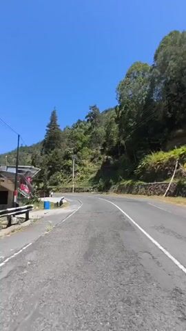 The video shows a road curving through a forested mountainside under a clear blue sky. On the left side of the road, there is a building with a red and white sign, and a blue trash can. Further down the road, there are a few people walking. The road is paved and has white lines marking the lanes. The trees on the right side of the road are dense and green. The camera appears to be moving forward, giving a first-person perspective of the journey.