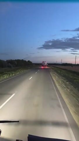 The video shows a dashcam view from a vehicle driving on a highway at dusk. The sky is a deep blue with hints of pink and orange from the setting sun. The road is asphalt with white lane markings, and there are green fields and trees on either side. 

Several vehicles are visible on the road. A dark-colored SUV is driving in front, followed by a larger truck. The SUV has what appear to be bike racks attached to the rear. The truck's taillights are also illuminated. The dashcam itself is visible at the bottom of the frame, along with parts of the vehicle's interior, including the windshield wipers and dashboard. The lighting suggests it is either early morning or late evening. 

The scene appears to be rural, likely in Kazakhstan as indicated by the location context. There are no people clearly visible in the video, but the presence of multiple vehicles suggests traffic. The overall mood is calm and serene, typical of a drive during twilight hours. No discernible text is visible in the footage.