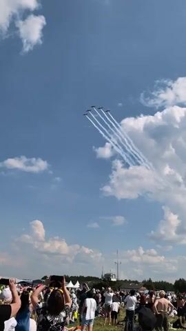 A flight of five small aircraft flies in formation across a clear blue sky with scattered white clouds. The planes are leaving white contrails that form parallel lines, fanning out as they move away from the viewer. Below the sky, a crowd of people is gathered, mostly facing upwards to watch the aerial display. The people are varied in age and attire, with some holding up cameras or phones to record the event. The overall scene suggests an airshow or similar outdoor public event.